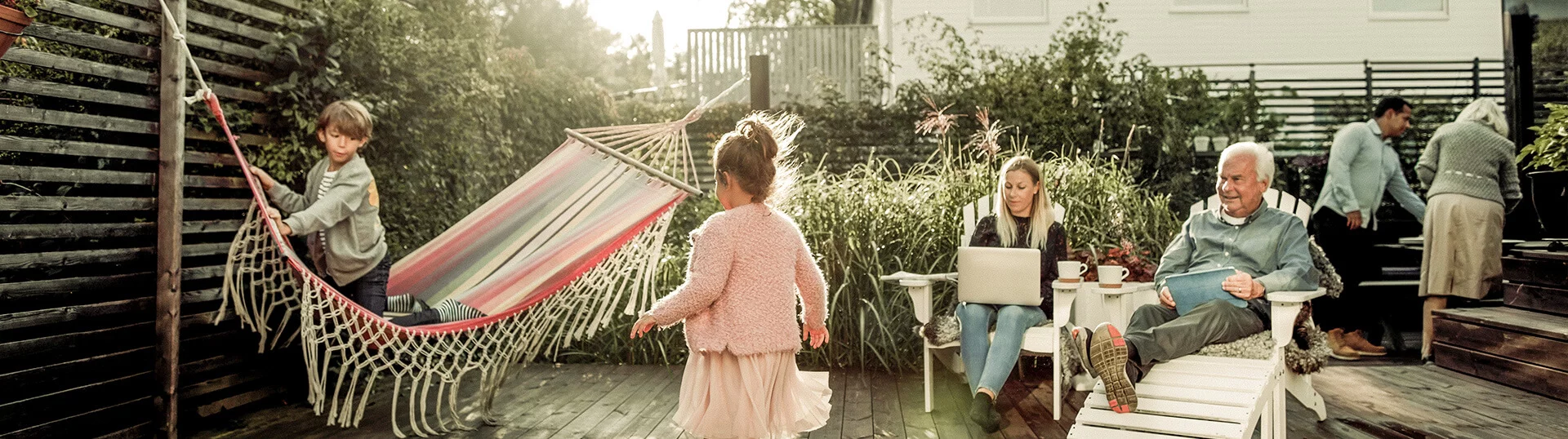 Scène de jardin avec un hamac, un enfant jouant et des adultes assis, ambiance ensoleillée et détendue.